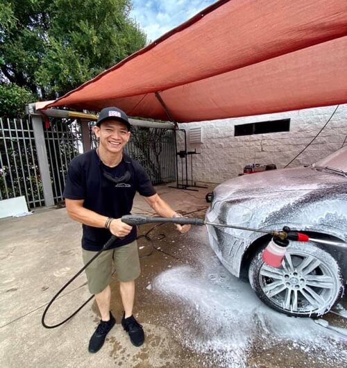 EurAuto Shop in Plano Tx, team member pictured smiling on a sunny day, in uniform t shirt and hat with shorts, is washing a vehicle at the shop under a shade canopy tent that is red