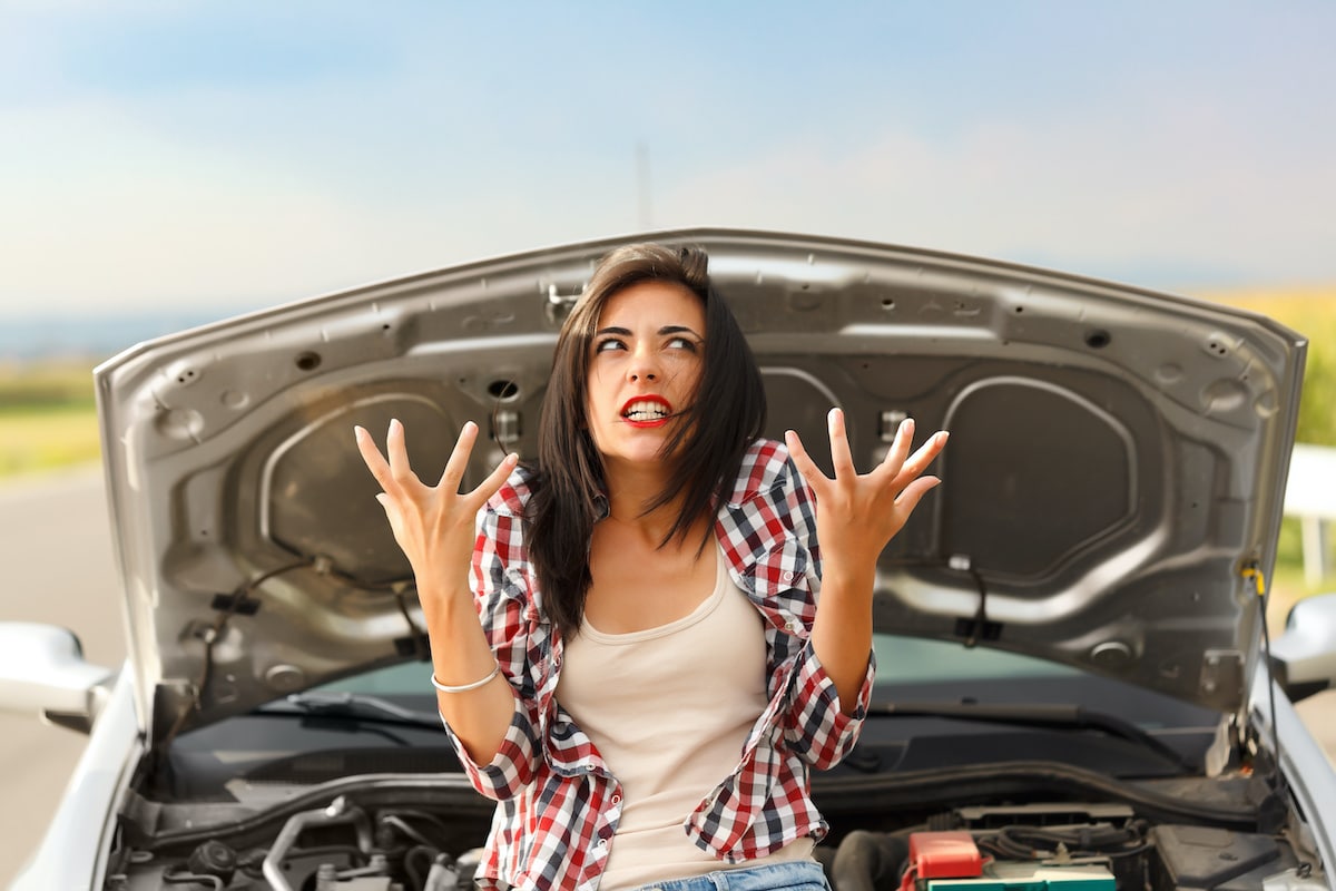 woman looking frustrated with hands up in air while sitting on hood of car engine with hood open on side of road