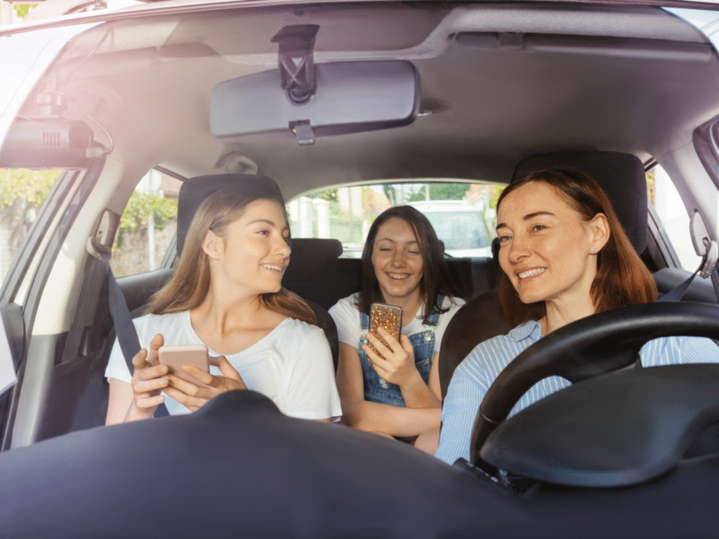 Summer road trip preparation by EurAuto Shop, Plano, TX. Image shows a mother and her teenage daughters inside a car, smiling and using their smartphones, emphasizing the importance of a well-maintained A/C system for a comfortable and enjoyable journey.