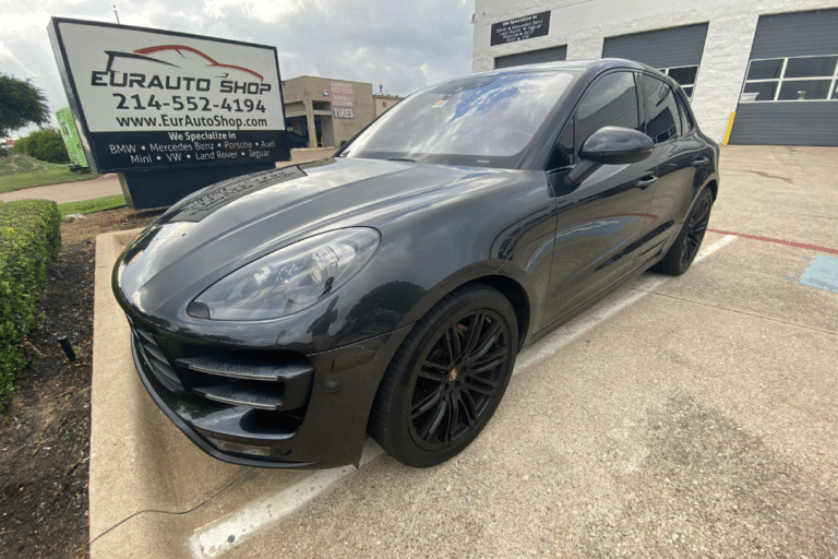 Porsche Macan, Porsche SUV In Plano TX At EurAuto Shop. Image of a dark gray Porsche Macan SUV parked in a concrete lot in front of a EurAuto Shop sign and garage facility.